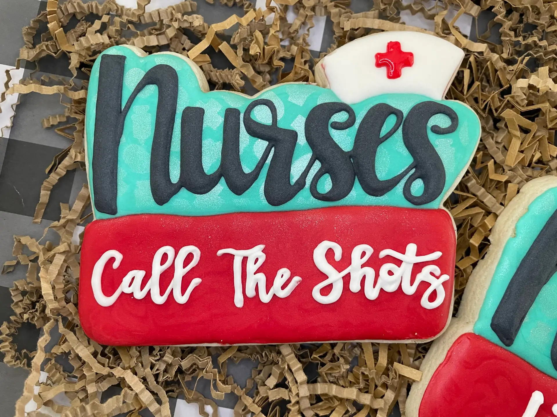 Large nurse-themed custom cookie with teal, red, and white royal icing and the phrase “Call The Shots”